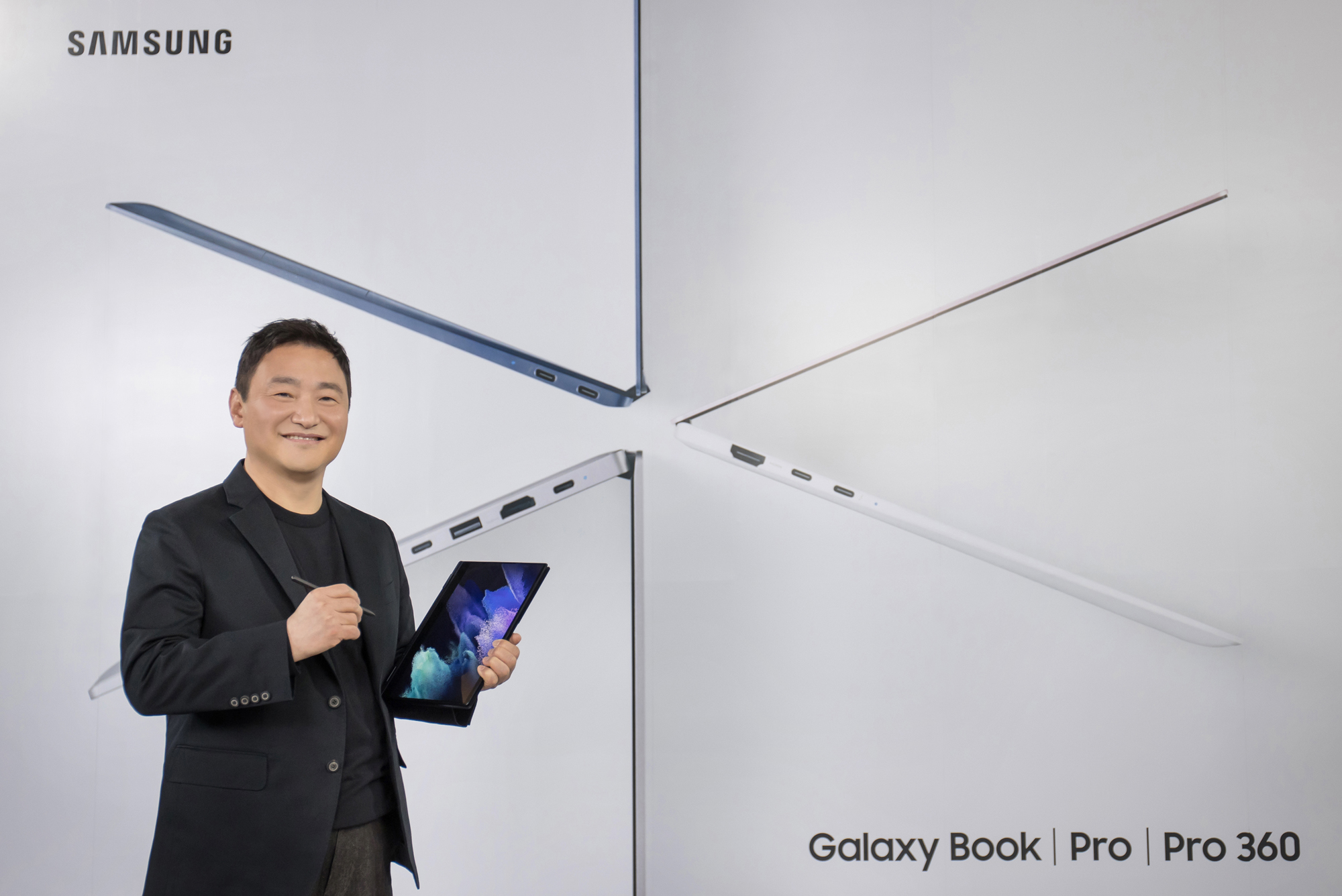 A man dressed wearing a dark suit jacket holding a Galaxy Book Pro 360 with his left hand while holding a S Pen with his right standing in front of a key visual image of galaxy book galaxy book pro and galaxy book pro 360