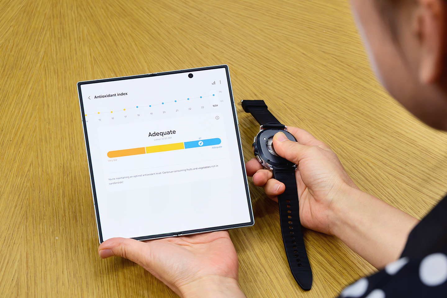 A person holds an unfolded Galaxy Fold7 and a Galaxy Watch8, pressing their thumb against the back of the watch. The phone screen reveals the Antioxidant Index score.