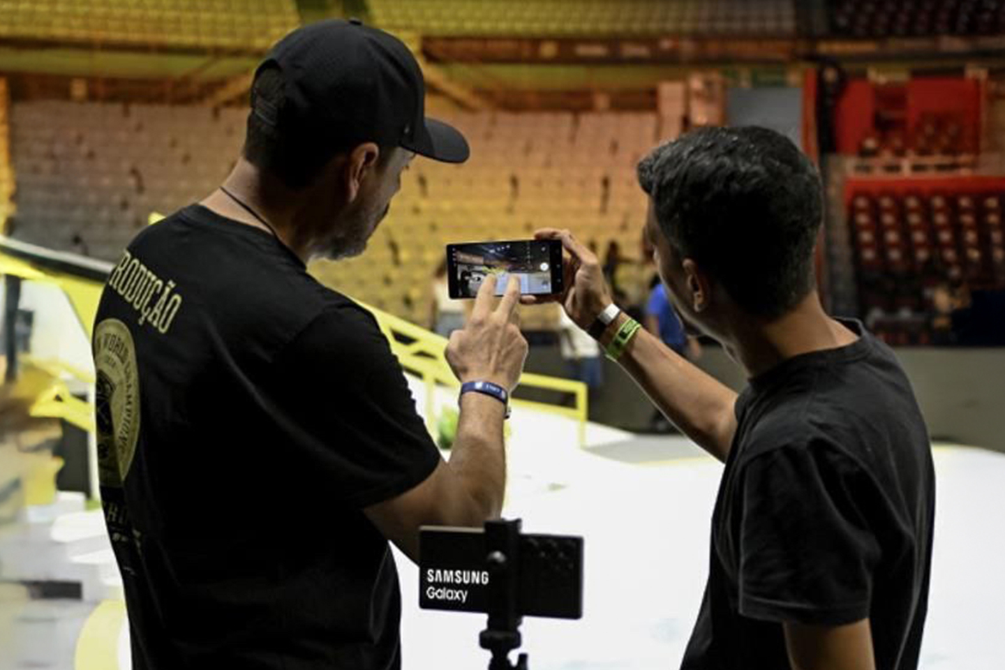 Two men stand at the SLS Super Crown World Championship stage. One holds a Galaxy S25 pointed toward the stage while the other adjusts its camera. A second Galaxy S25 is mounted nearby.